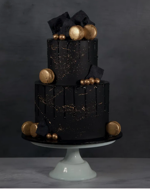 A two-tiered black drip cake decorated with gold accents, black frosting, and edible golden balls, presented on a white cake stand. Available for next-day delivery.