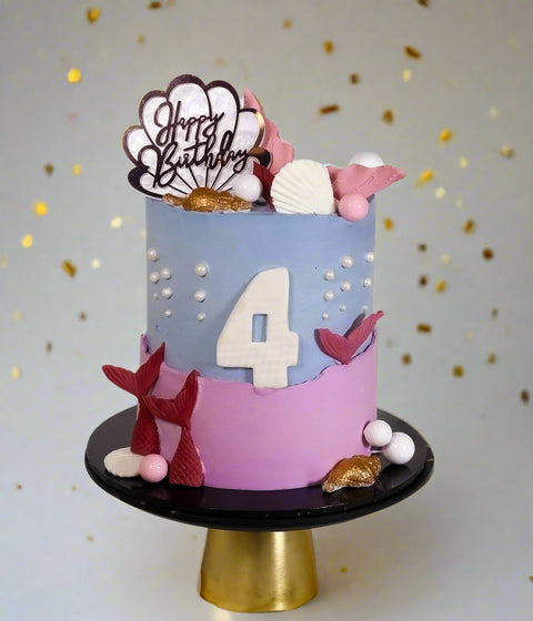 A decorated birthday cake with mermaid-themed elements, including pink, lilac, white, and gold tones, mermaid tails, sea shells, and the age
