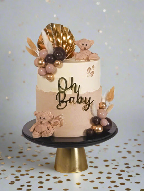 Round cake with beige and brown layers, decorated with gold accents, teddy bear figures, and the phrase 'Oh Baby' on the cake, presented on a gold stand.