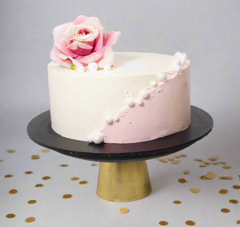 Decorative cake with pink and white frosting, rose, and pearls on a black and gold stand.